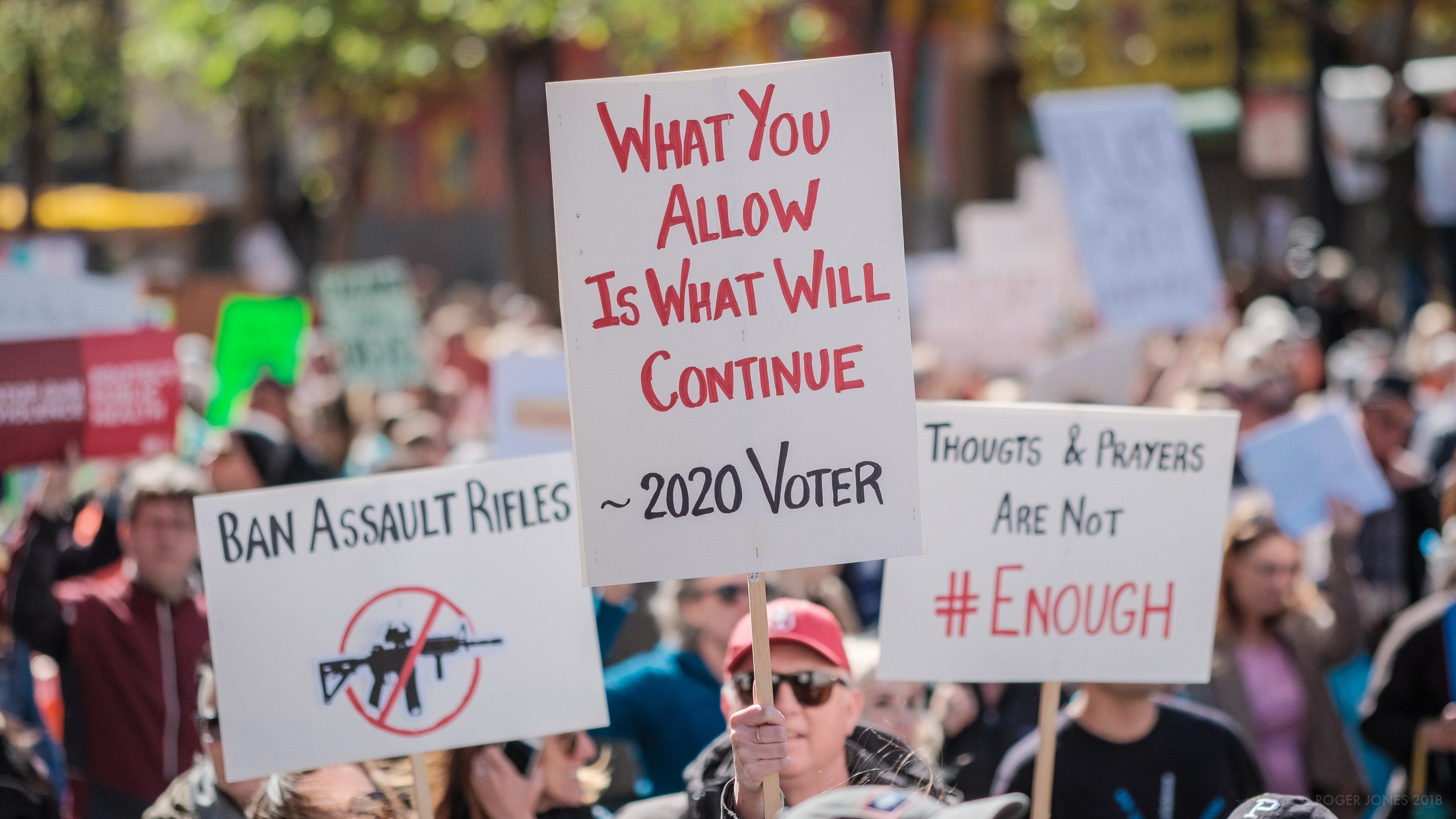 The image shows a crowd of people holding signs in front of a building, with trees in the...
