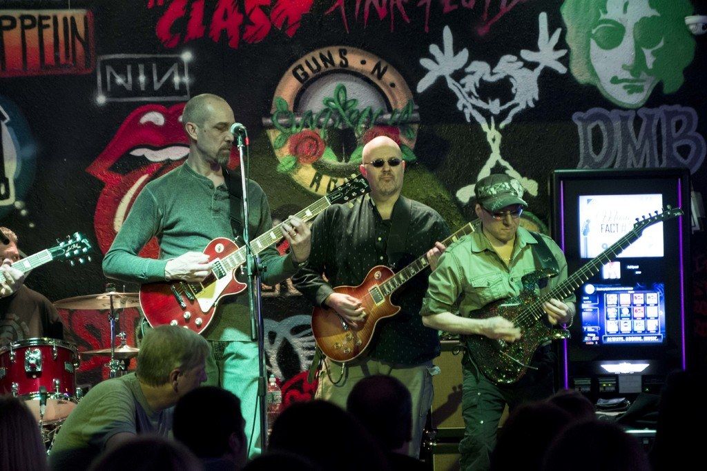 This image is clicked in a concert. There are three men in this image. At the bottom, there is a...