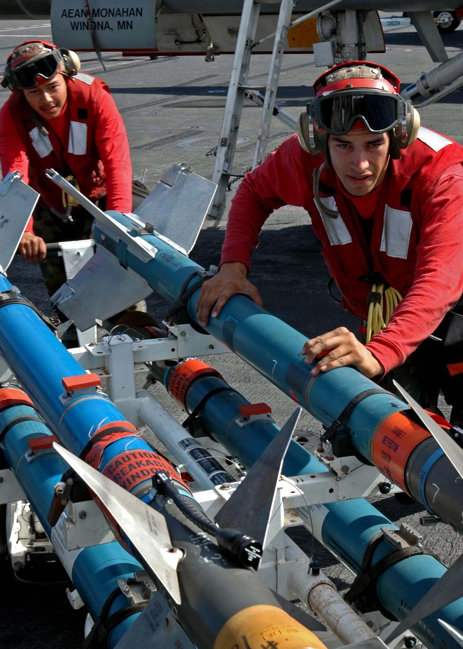 The image shows two men in red uniforms working on a missile on an aircraft carrier. They are...