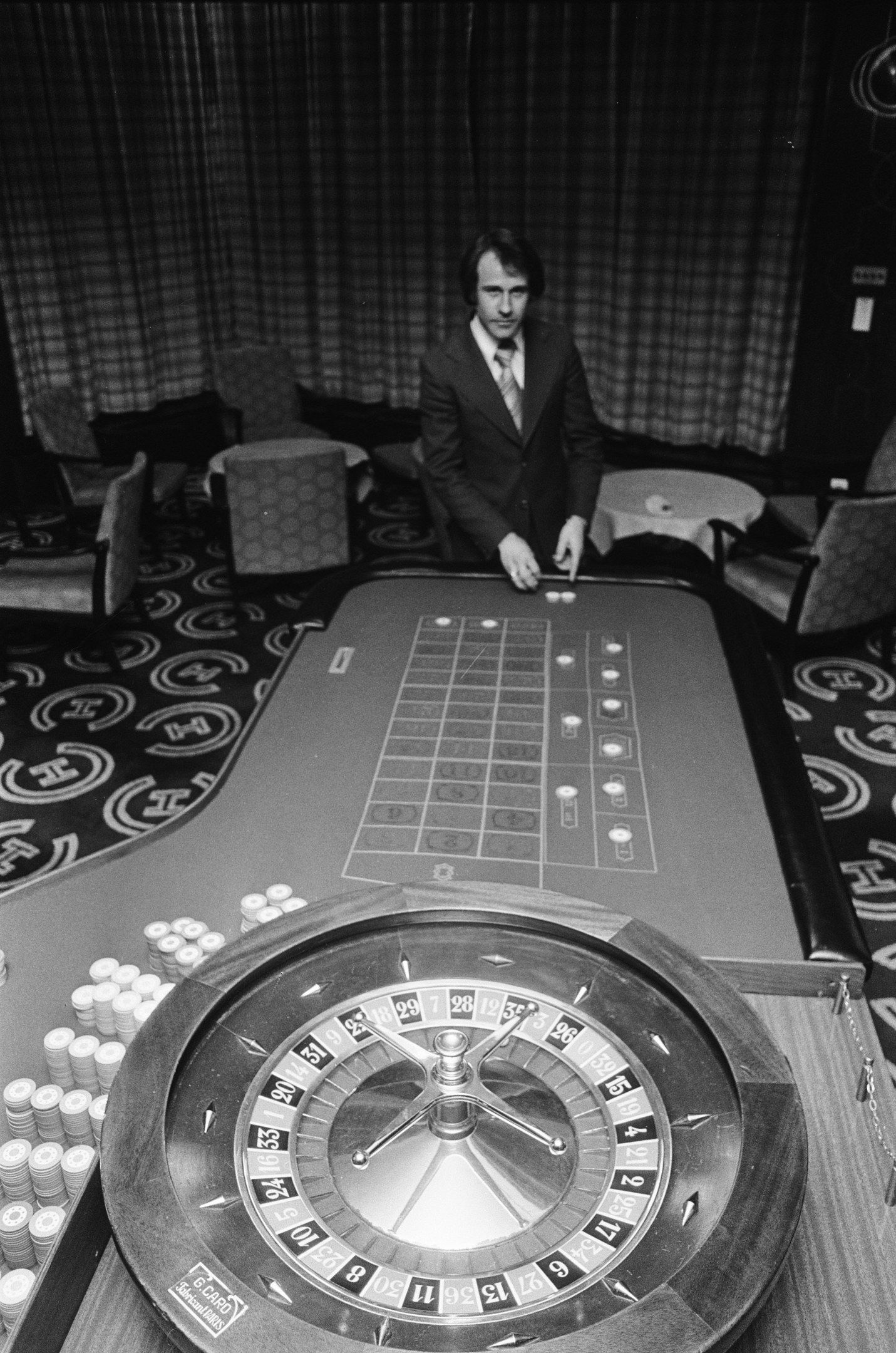 The image shows a man in a suit sitting at a roulette table in a casino. He is surrounded by chairs...