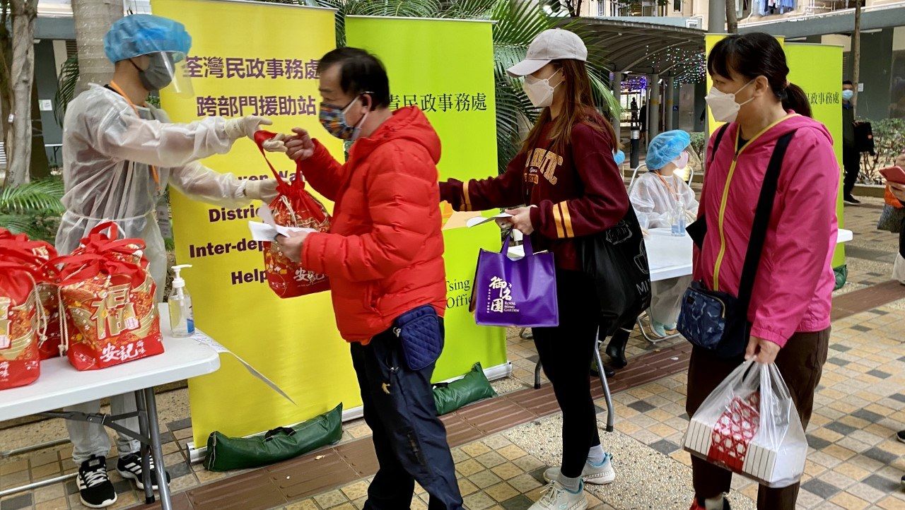 The image shows a group of people standing around a table with bags of food, wearing masks and...