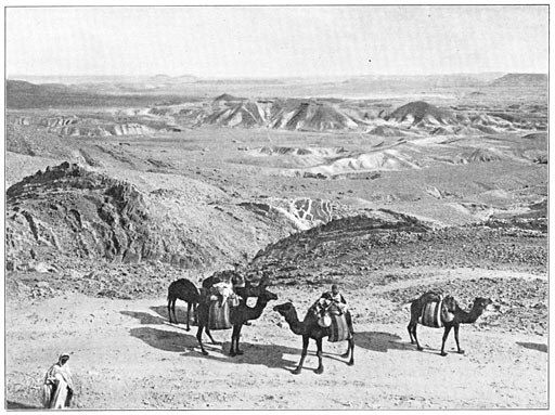 The image shows a group of people riding on the backs of camels in the desert, with hills in the...