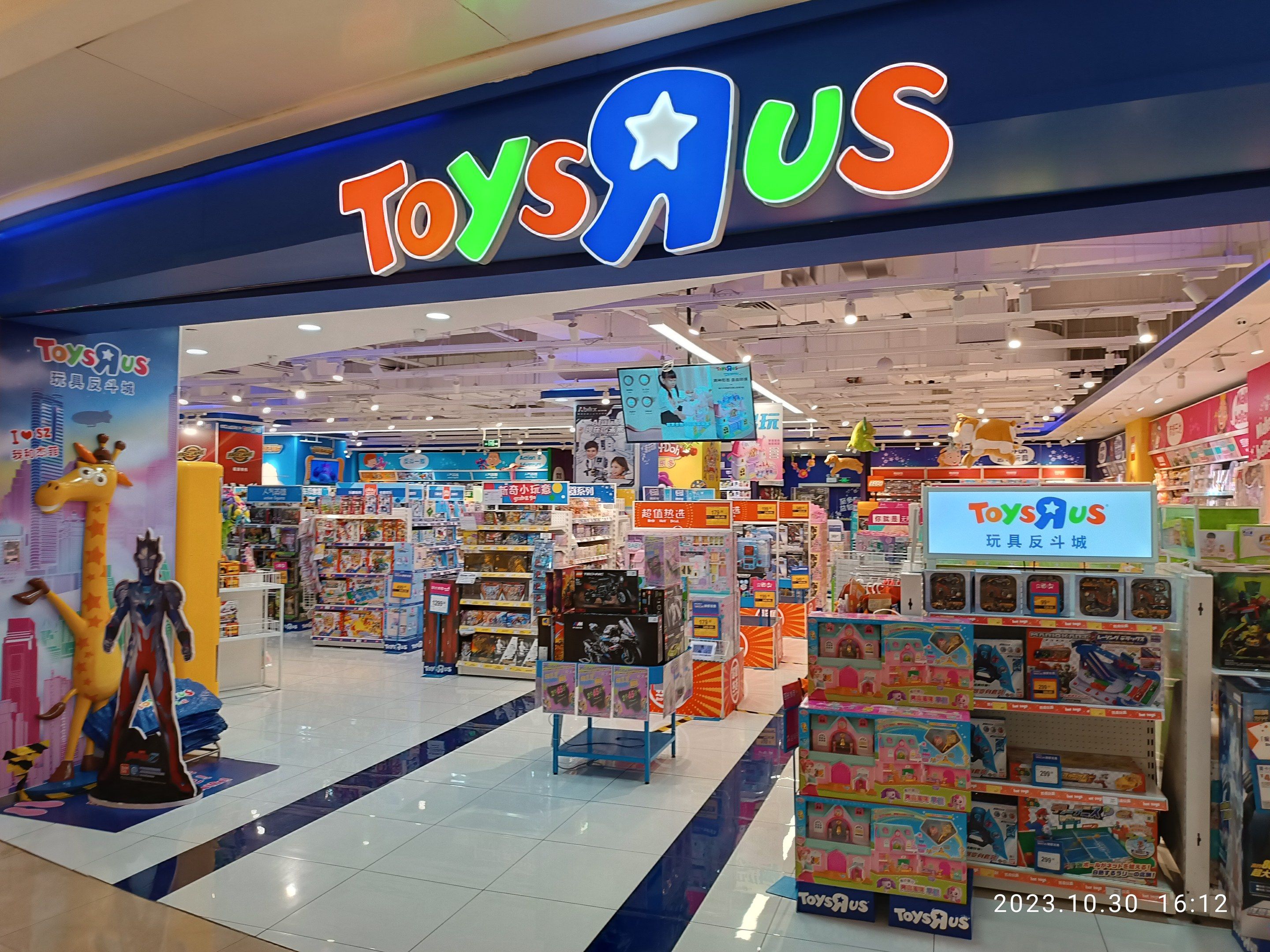 The image shows a Toys R Us store in a shopping mall, with shelves filled with toys and other...