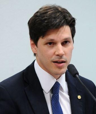 The image shows a man in a suit and tie speaking into a microphone in front of a wall. He appears...