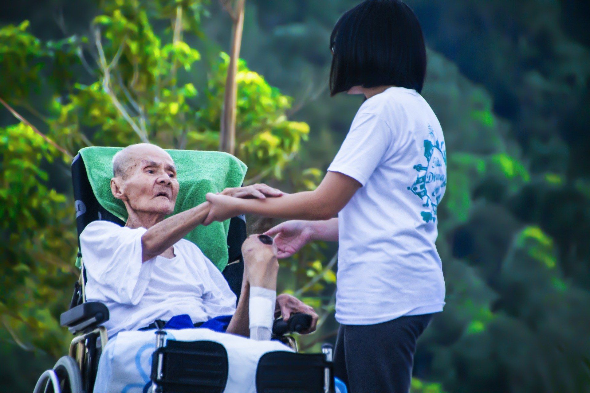 The image shows an elderly man in a wheelchair being assisted by a woman wearing a white t-shirt...