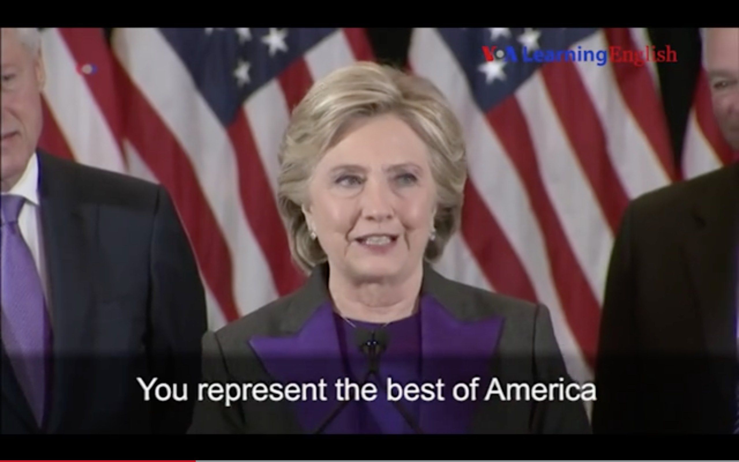 The image shows Hillary Clinton speaking at a podium in front of an American flag, with two men...