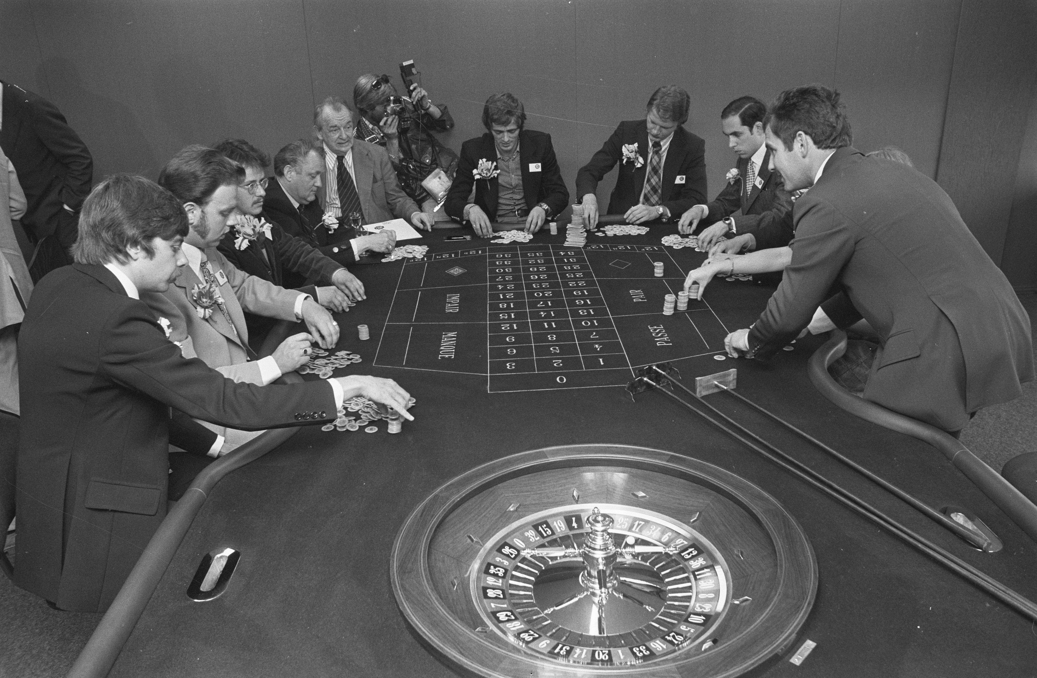 The image shows a group of men sitting around a roulette table in a casino. They are wearing...