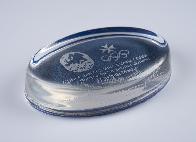 The image shows a clear glass award with a blue and white logo on it, sitting on a white surface....