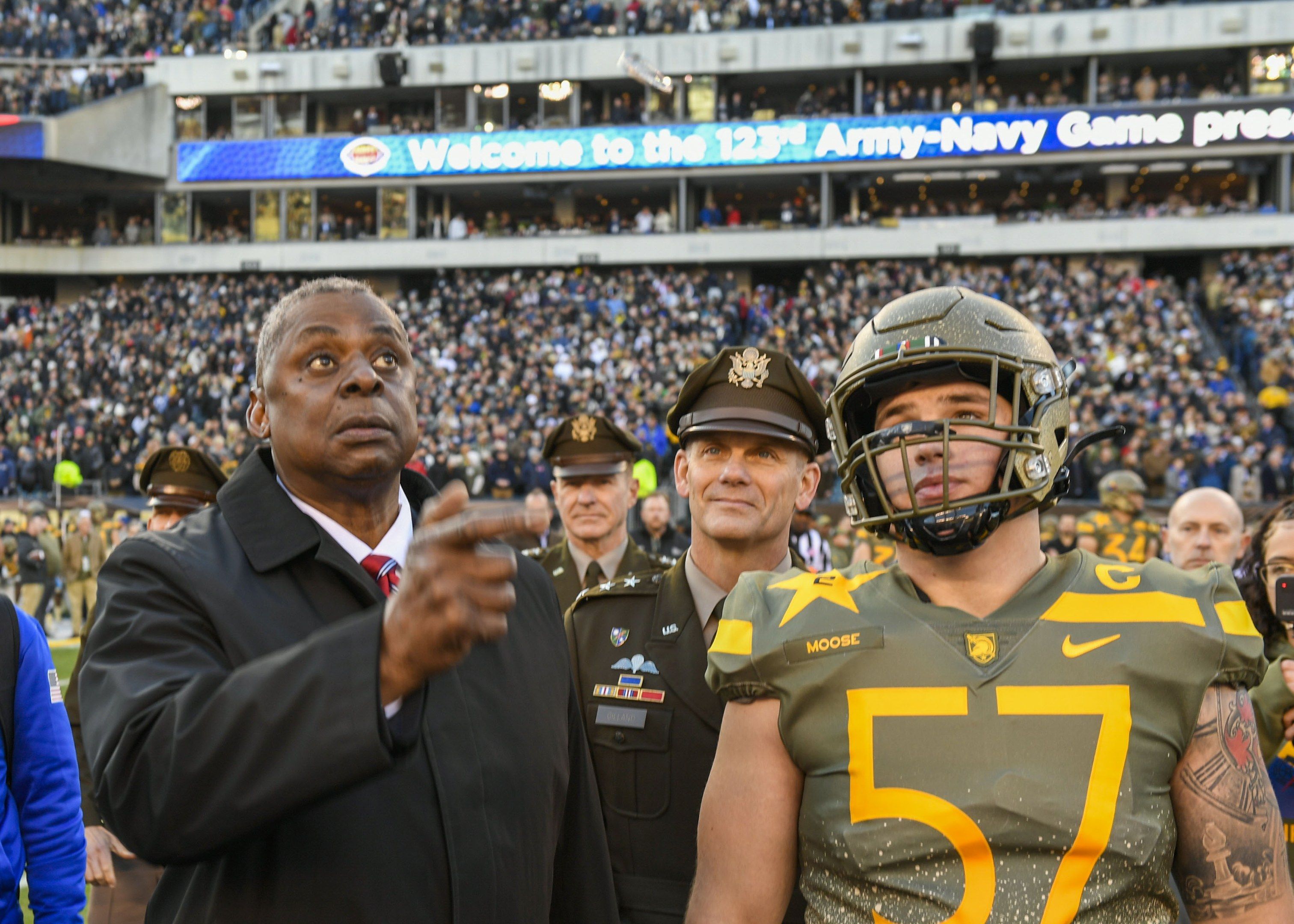 The image shows a man in a suit and tie standing next to a football player wearing a helmet,...