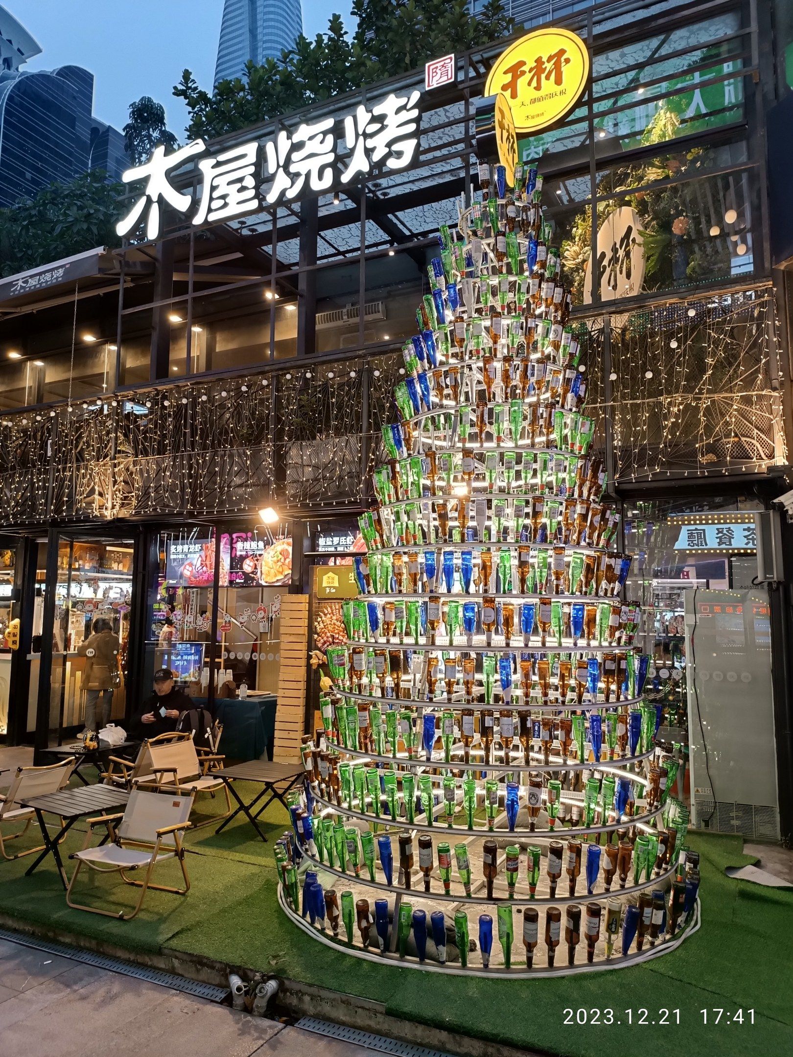 The image shows a festive Christmas tree made out of beer bottles in the middle of a city street....