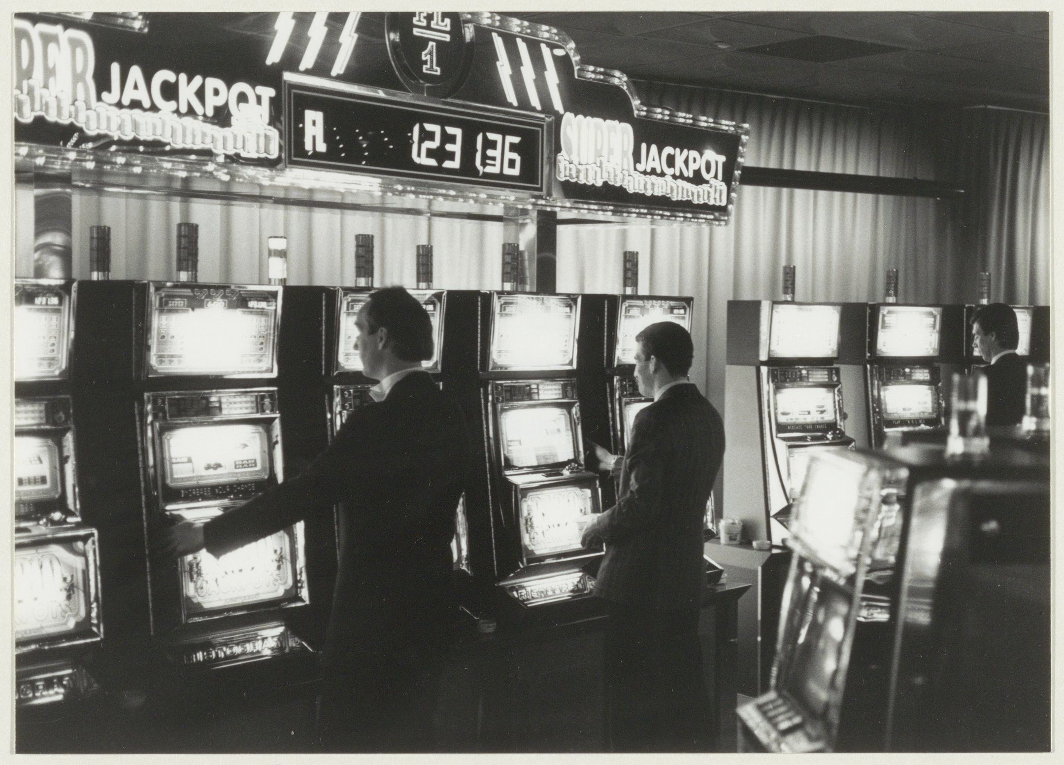 The image shows a black and white photo of two men playing slot machines in a casino. The men are...