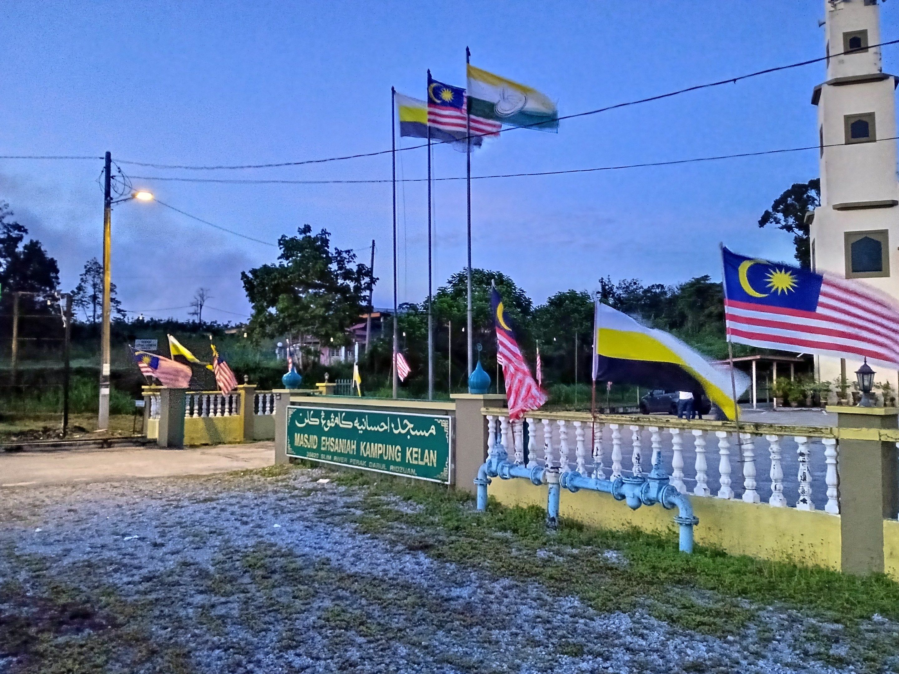 The image shows the entrance to a mosque with Malaysian flags flying in front of it. There is a...
