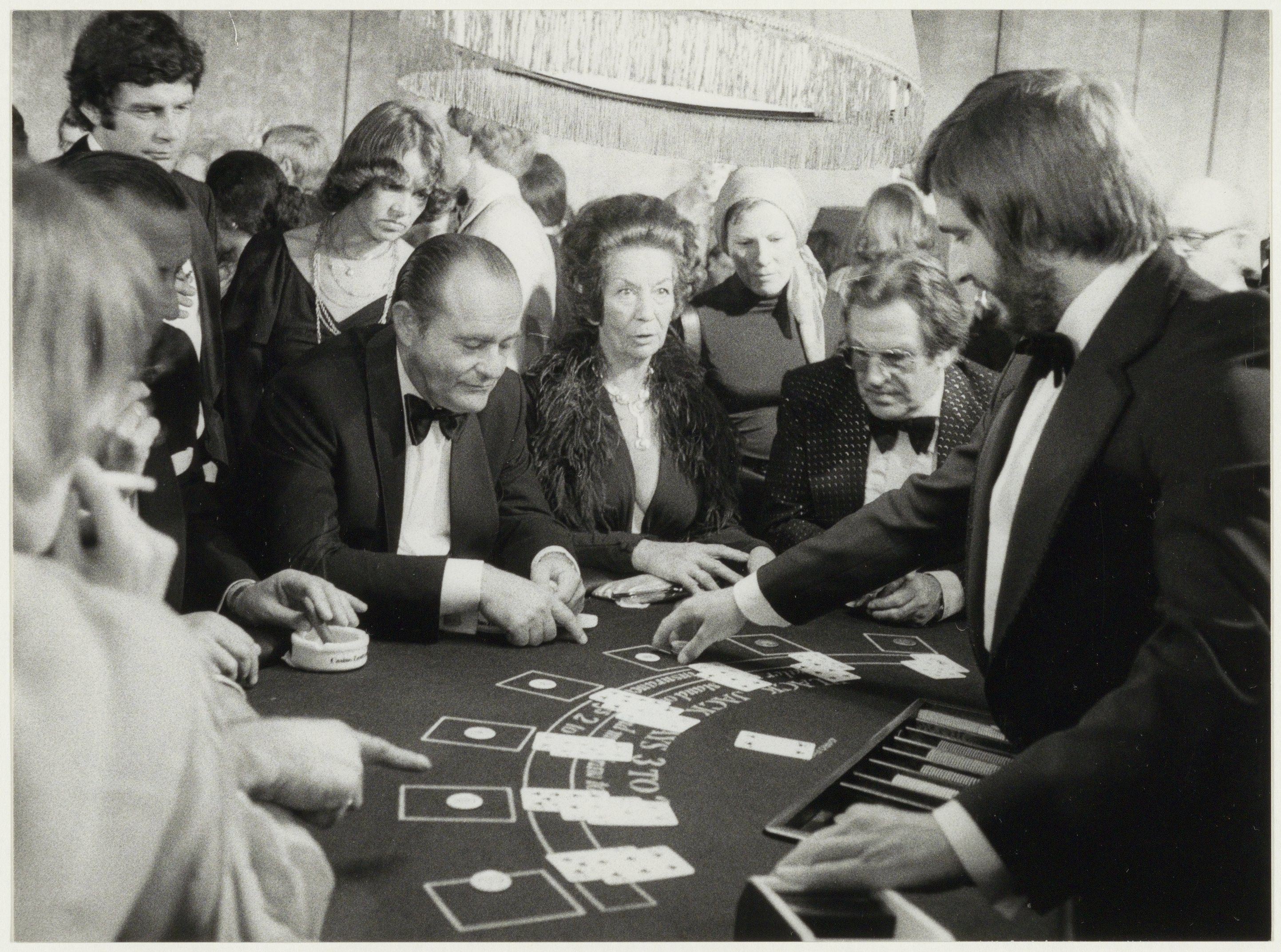 The image shows a group of people sitting around a blackjack table, with cards spread out in front...