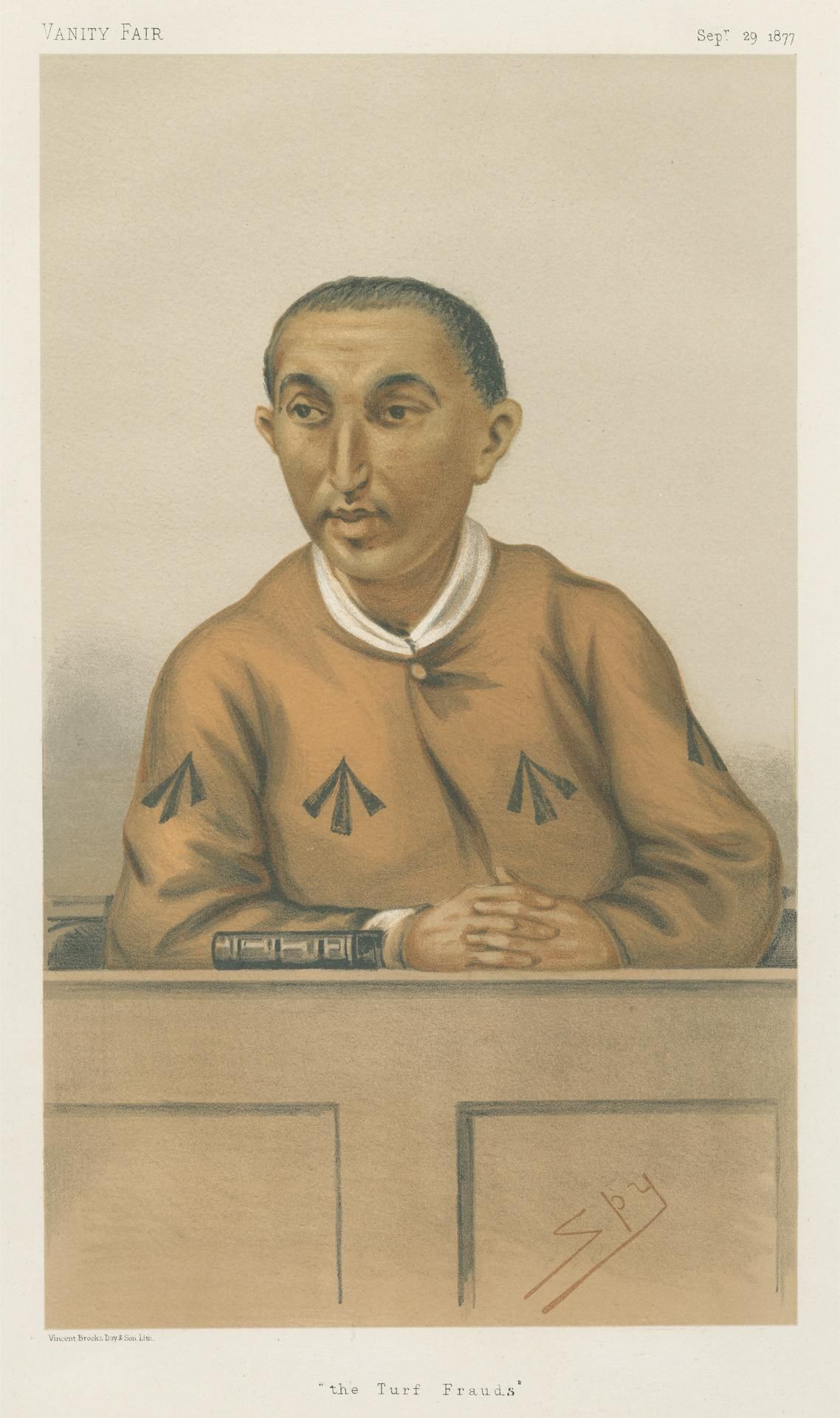 The image shows a man sitting at a table with his hands clasped in front of him. He is wearing a...