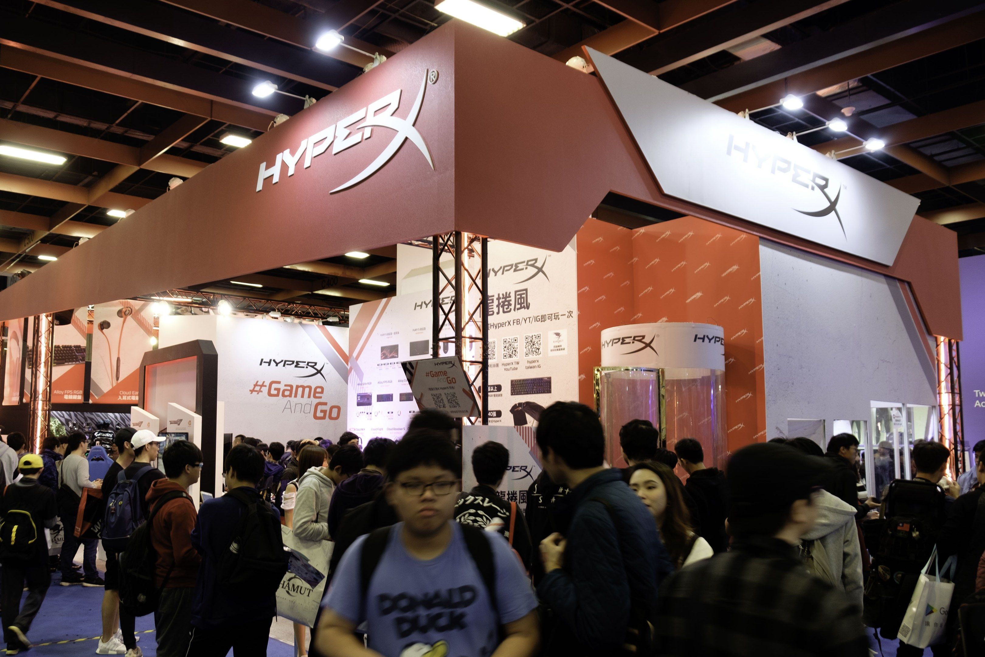 The image shows a large group of people standing in front of a booth at a gaming expo. Many of the...