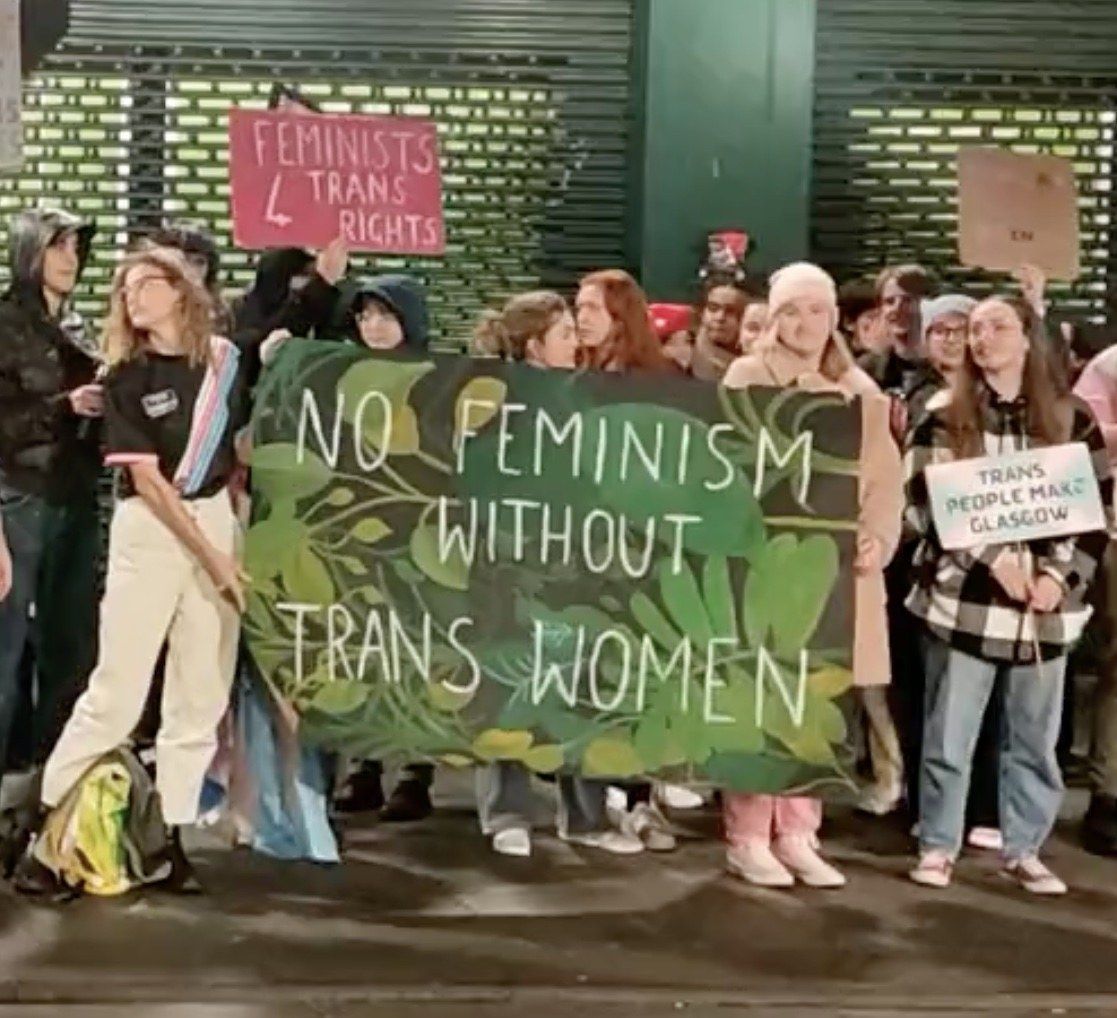 The image shows a group of people standing on the ground, holding a banner that reads "No Feminism...