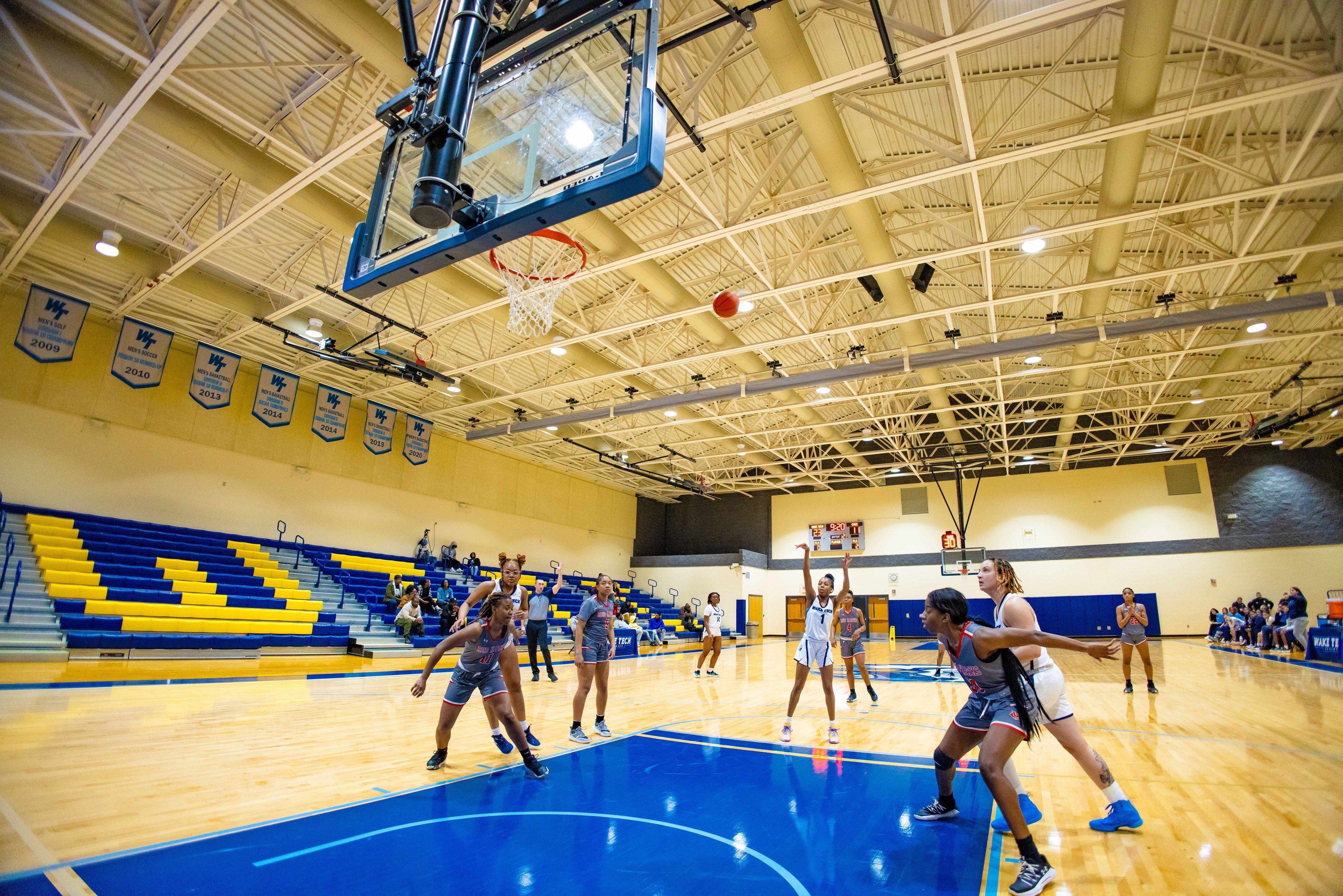 The image shows a group of women playing a game of basketball in a gymnasium. There are people...
