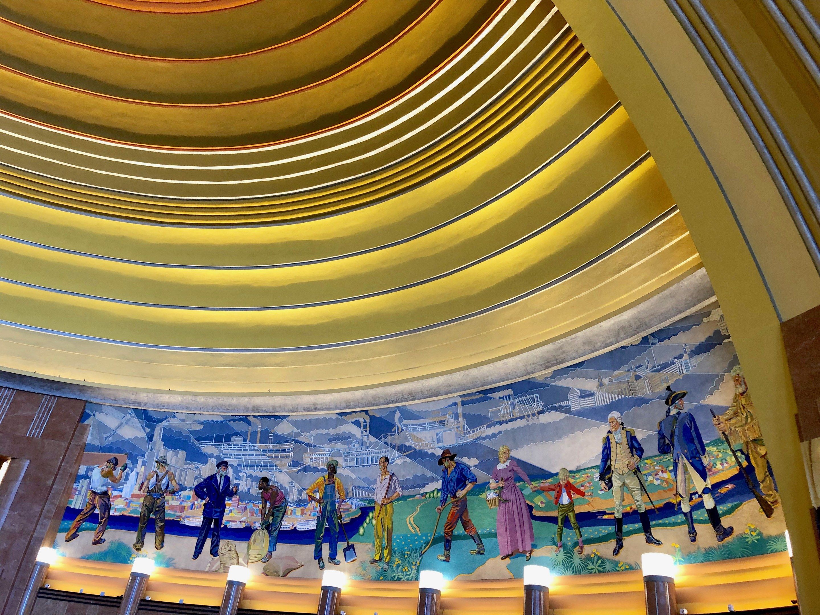 The image shows a large mural on the ceiling of a building, depicting people, buildings, and other...