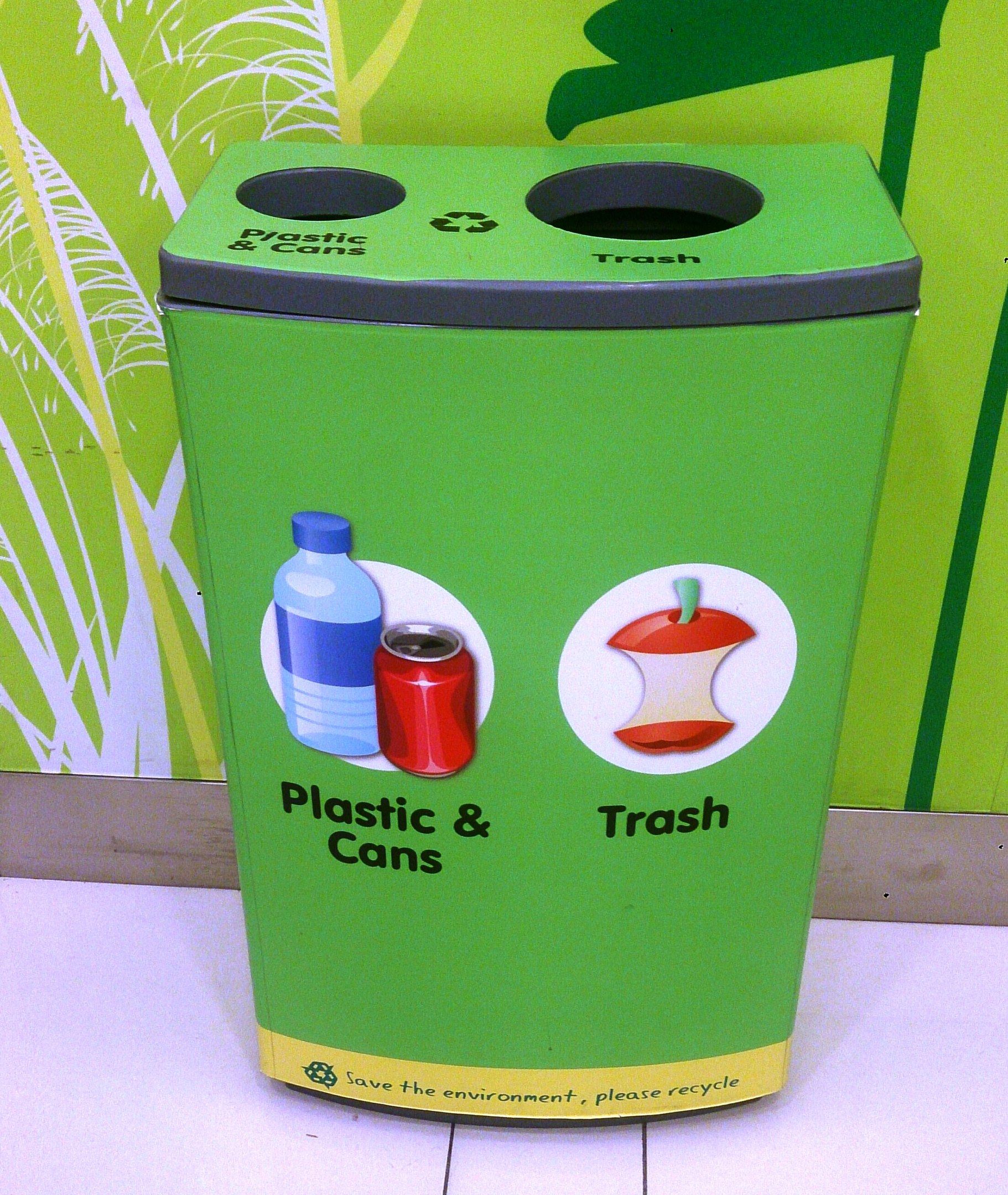 The image shows a green trash can sitting on top of a white floor, with a poster on the wall in the...