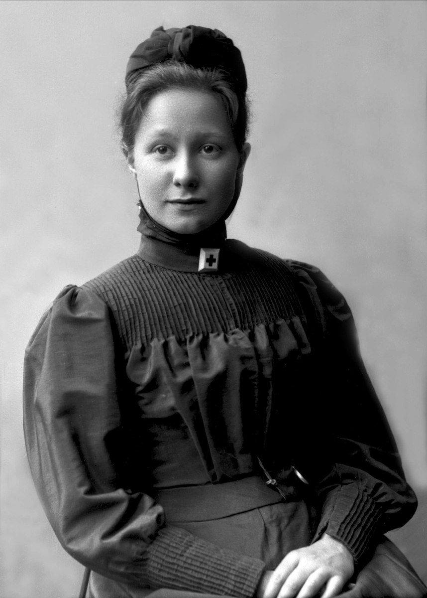 The image shows a black and white photo of a woman in a nurse's uniform, sitting on a chair. She...
