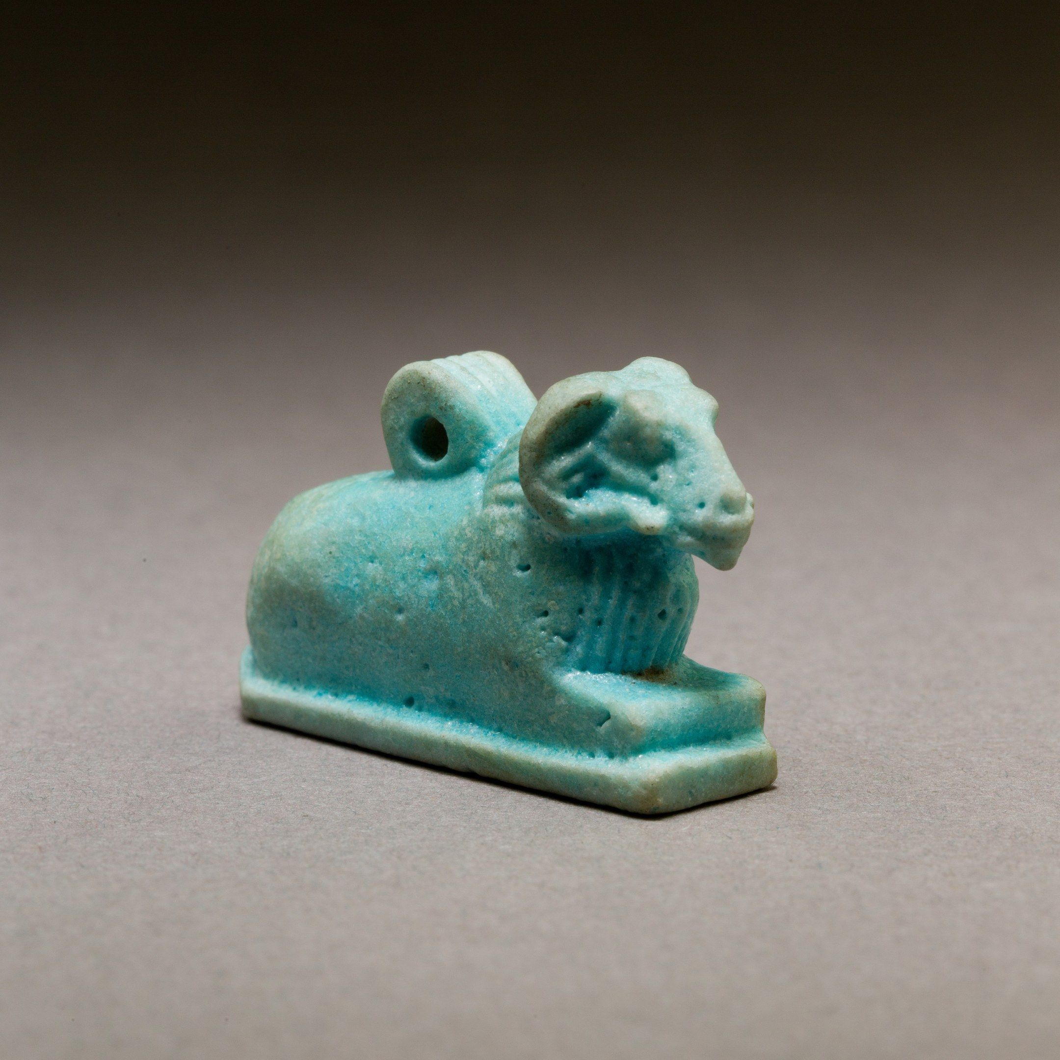 The image shows an ancient Egyptian faience amulet of a ram, which is blue in color and is placed...