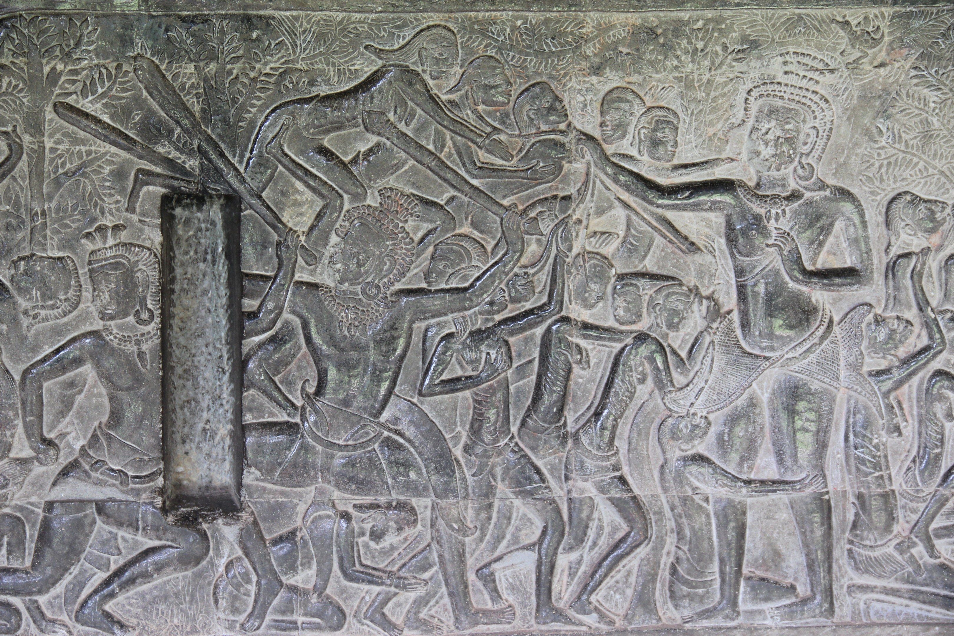 The image shows a stone carving on the wall of a temple in Angkor Thom, Cambodia depicting a scene...