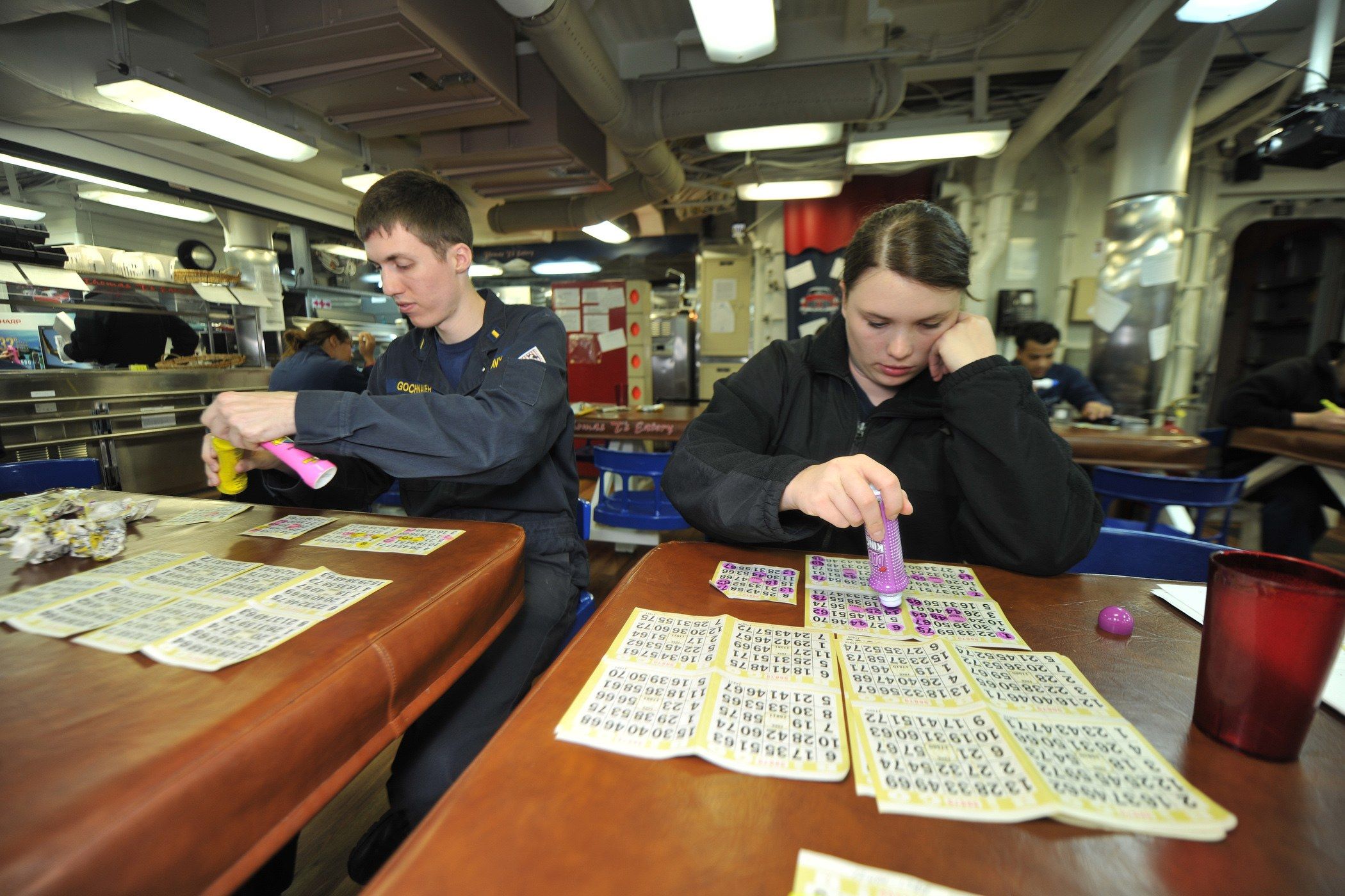 The image shows a man and woman sitting at a table playing a game of bingo. On the table there are...