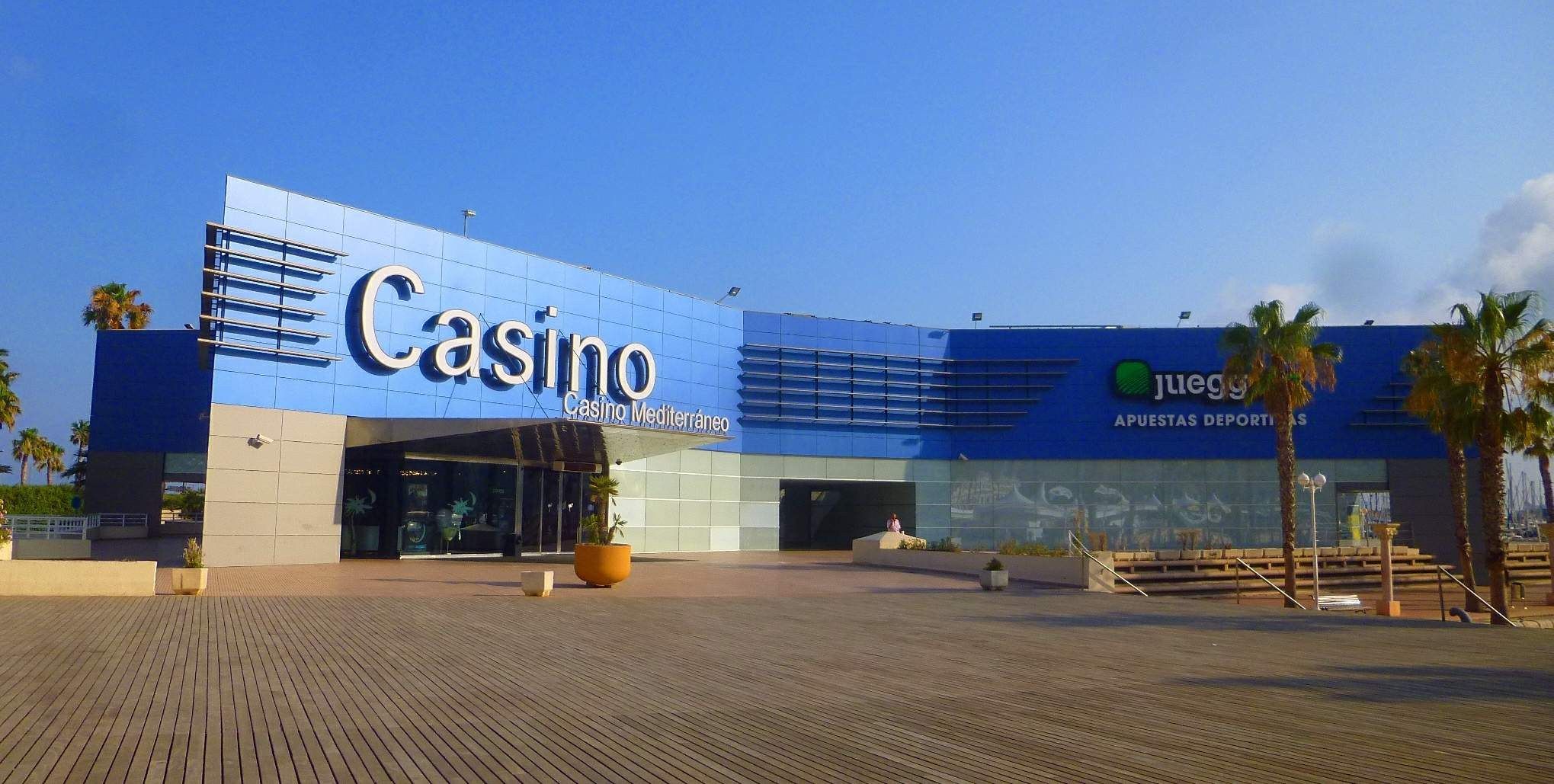 The image shows a casino building with palm trees in front of it, surrounded by plants in pots, a...