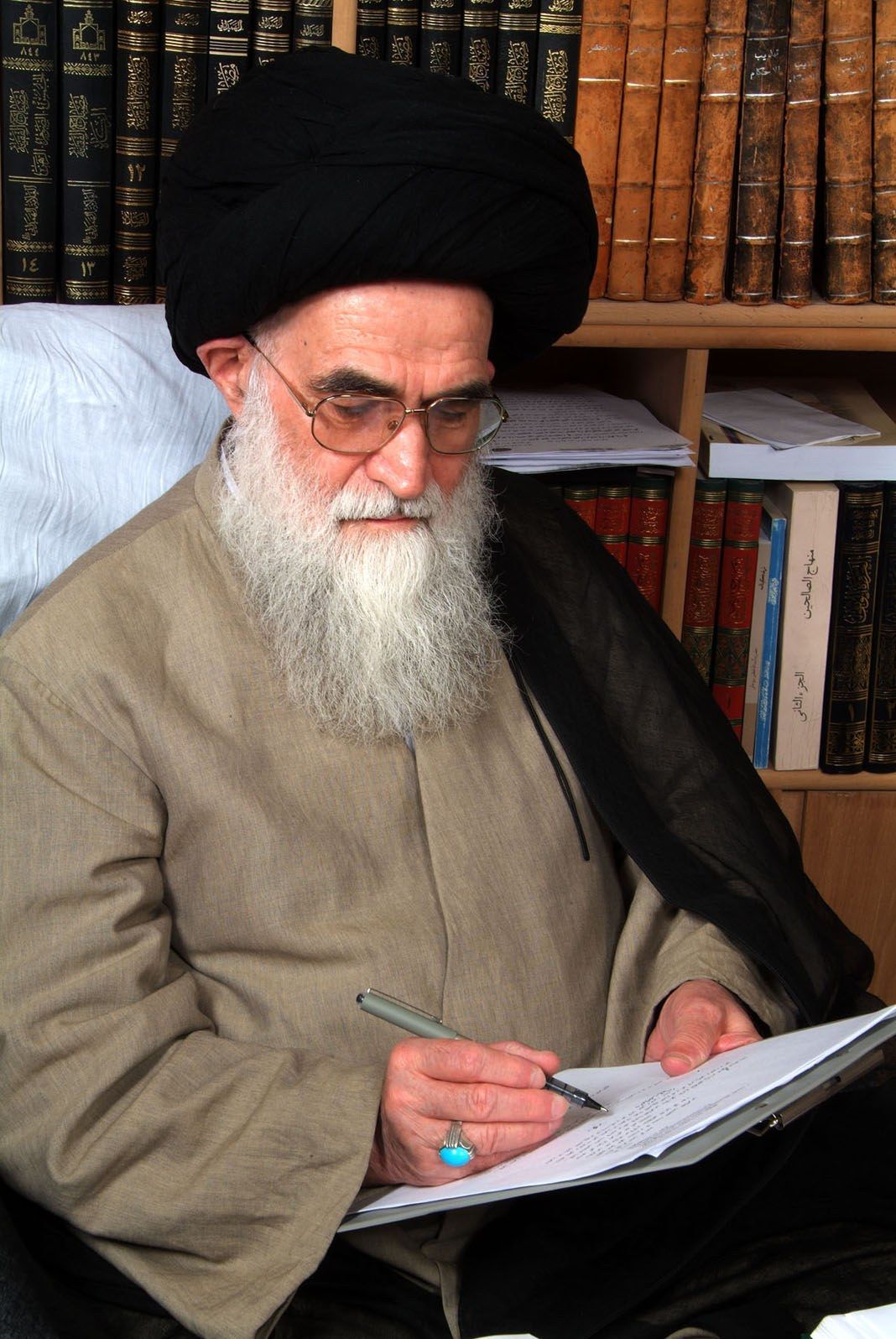 The image shows Ayatollah Ali Khamenei, the Supreme Leader of Iran, sitting on a chair and writing...