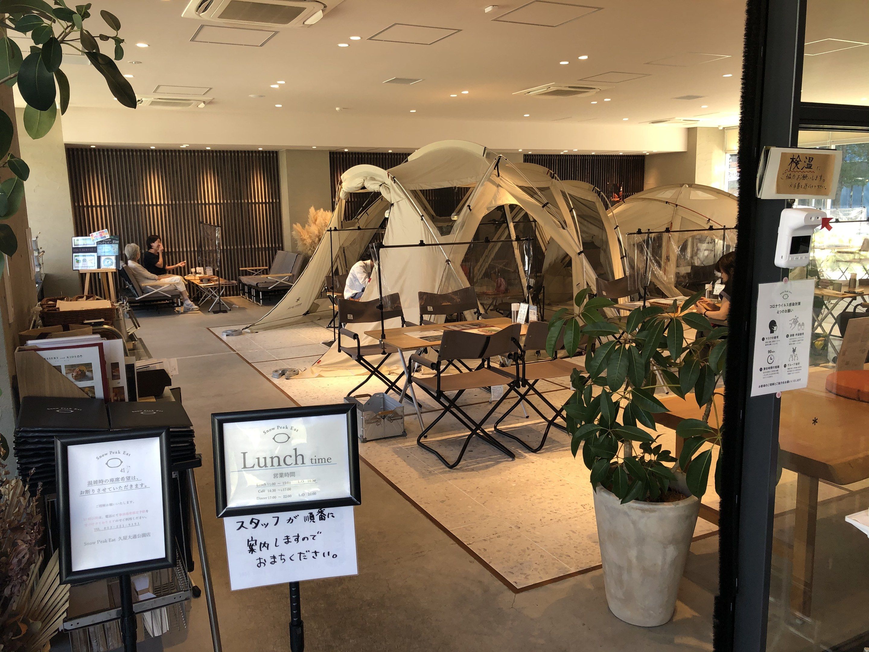 The image shows a restaurant with tables and chairs in front of a glass door, surrounded by plants,...