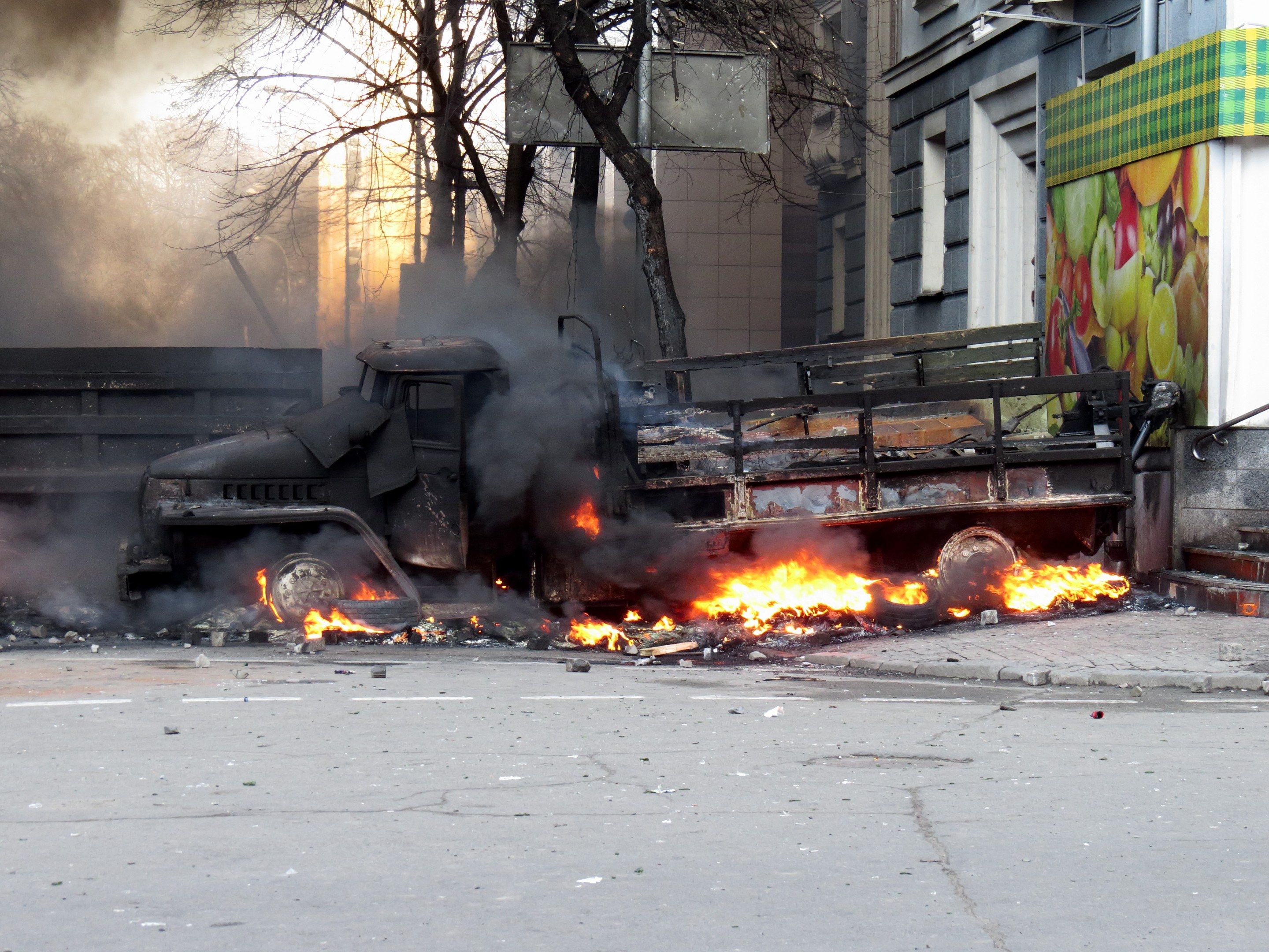 The image shows a truck engulfed in flames on the side of a street, with smoke billowing out of it....
