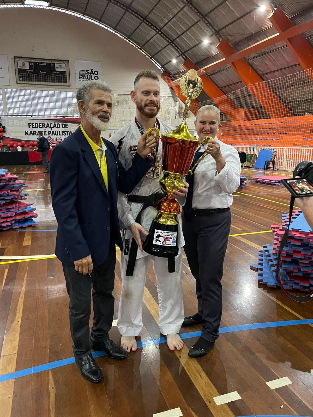 The image shows three men standing next to each other, proudly holding a trophy. On the right side...