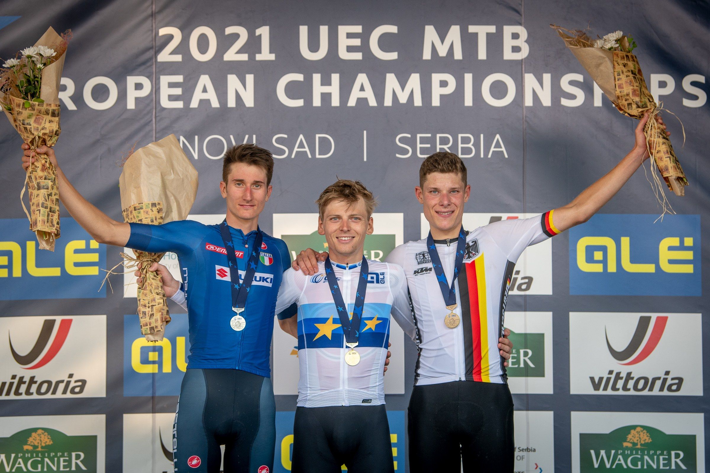The image shows three men standing on top of a podium, wearing medals and holding flower bouquets...