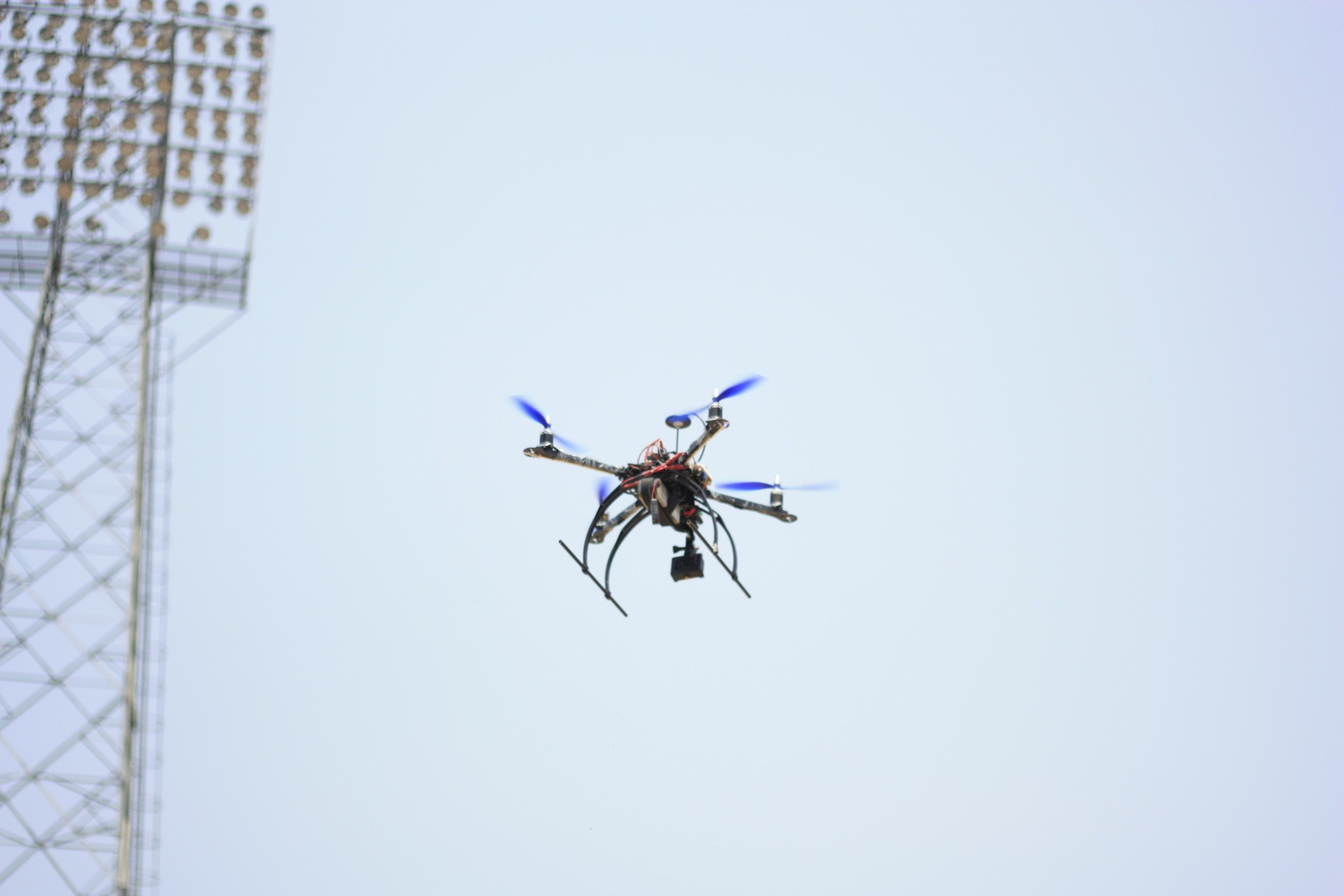 The image shows a drone flying in the air with a tower on the left side and a clear blue sky in the...