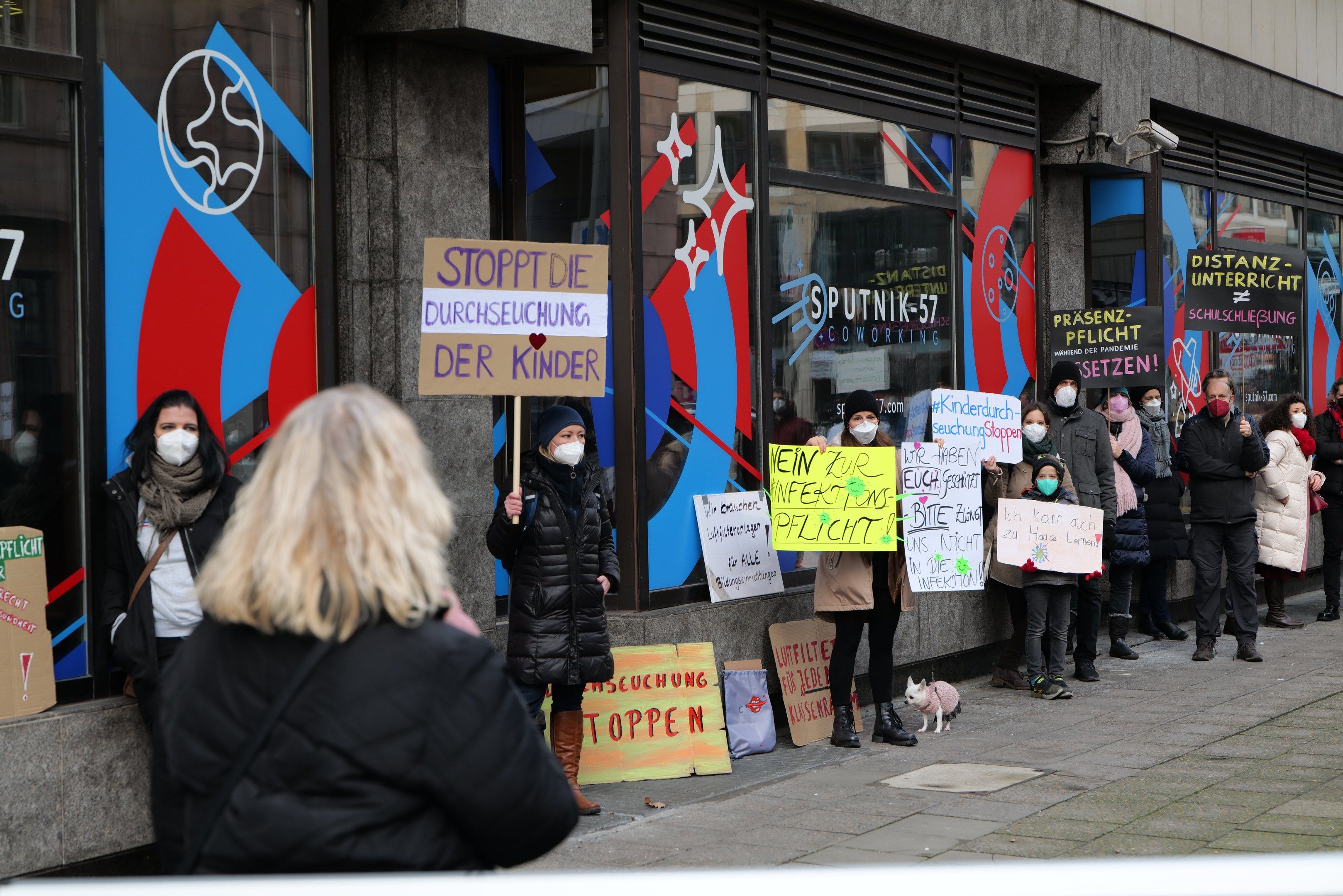 The image shows a group of people wearing masks and holding signs in front of a building with glass...