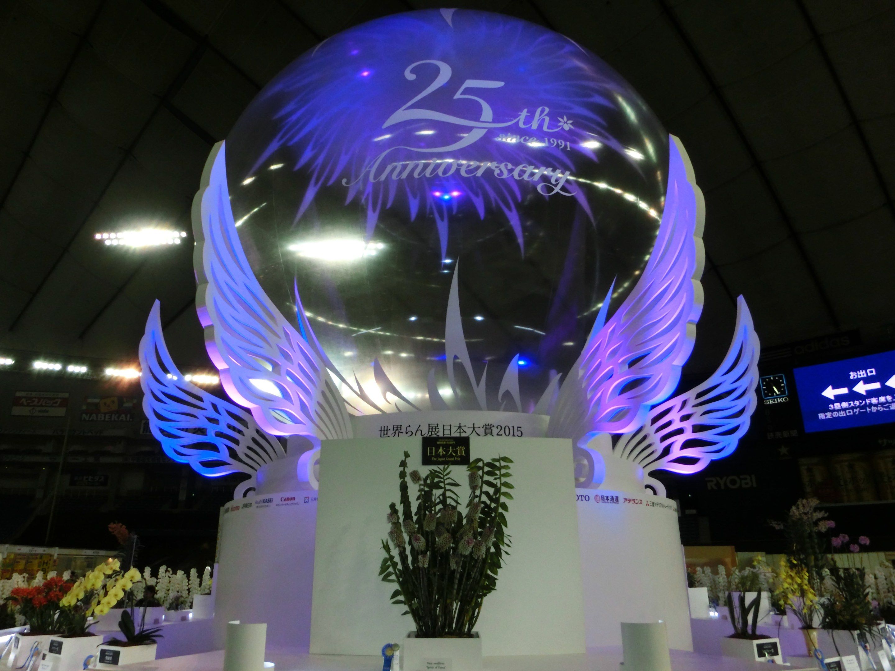 The image shows a large crystal ball with wings on top of it, surrounded by flower bouquets, name...