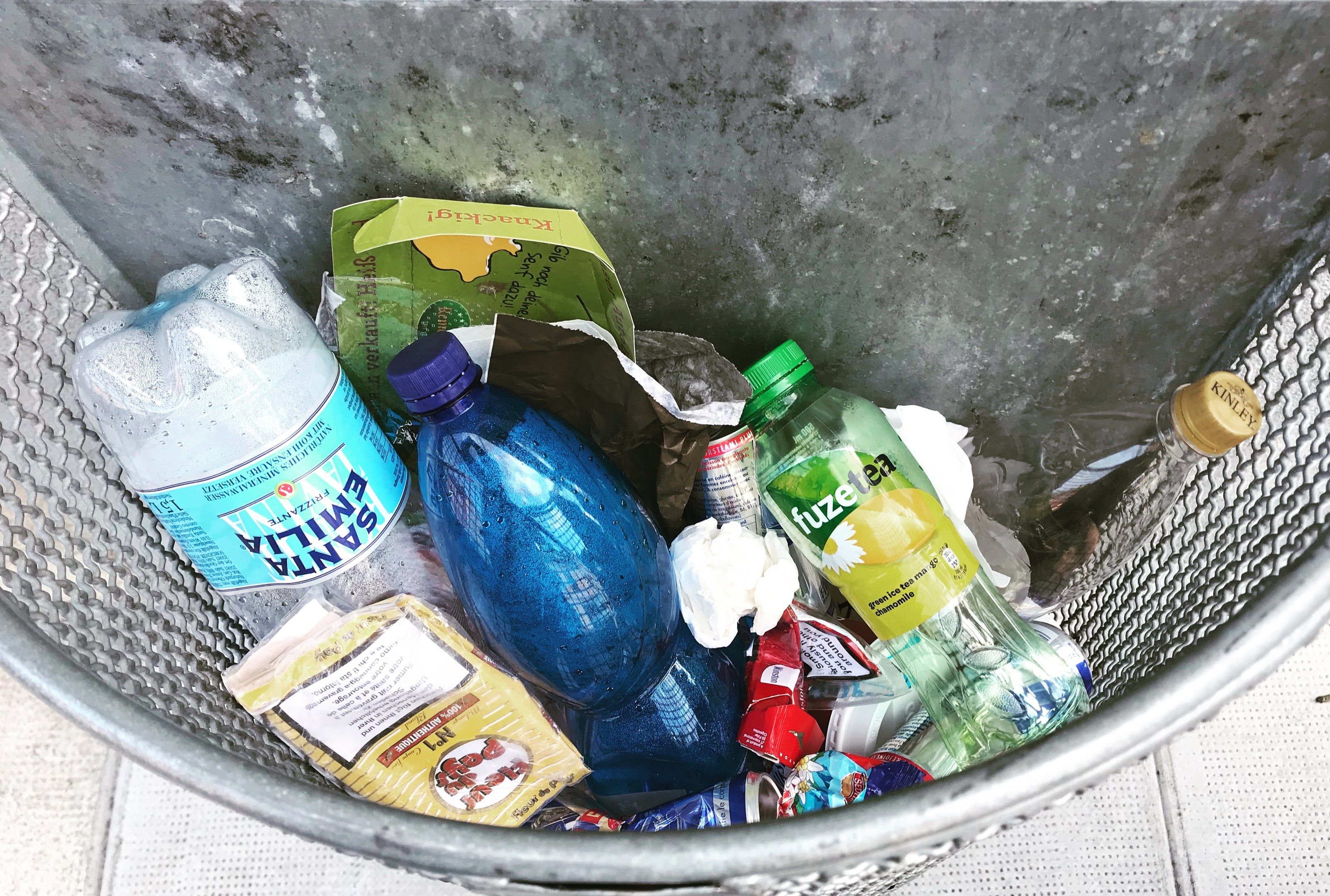 The image shows a metal trash can filled with a variety of plastic bottles, packets, and other...