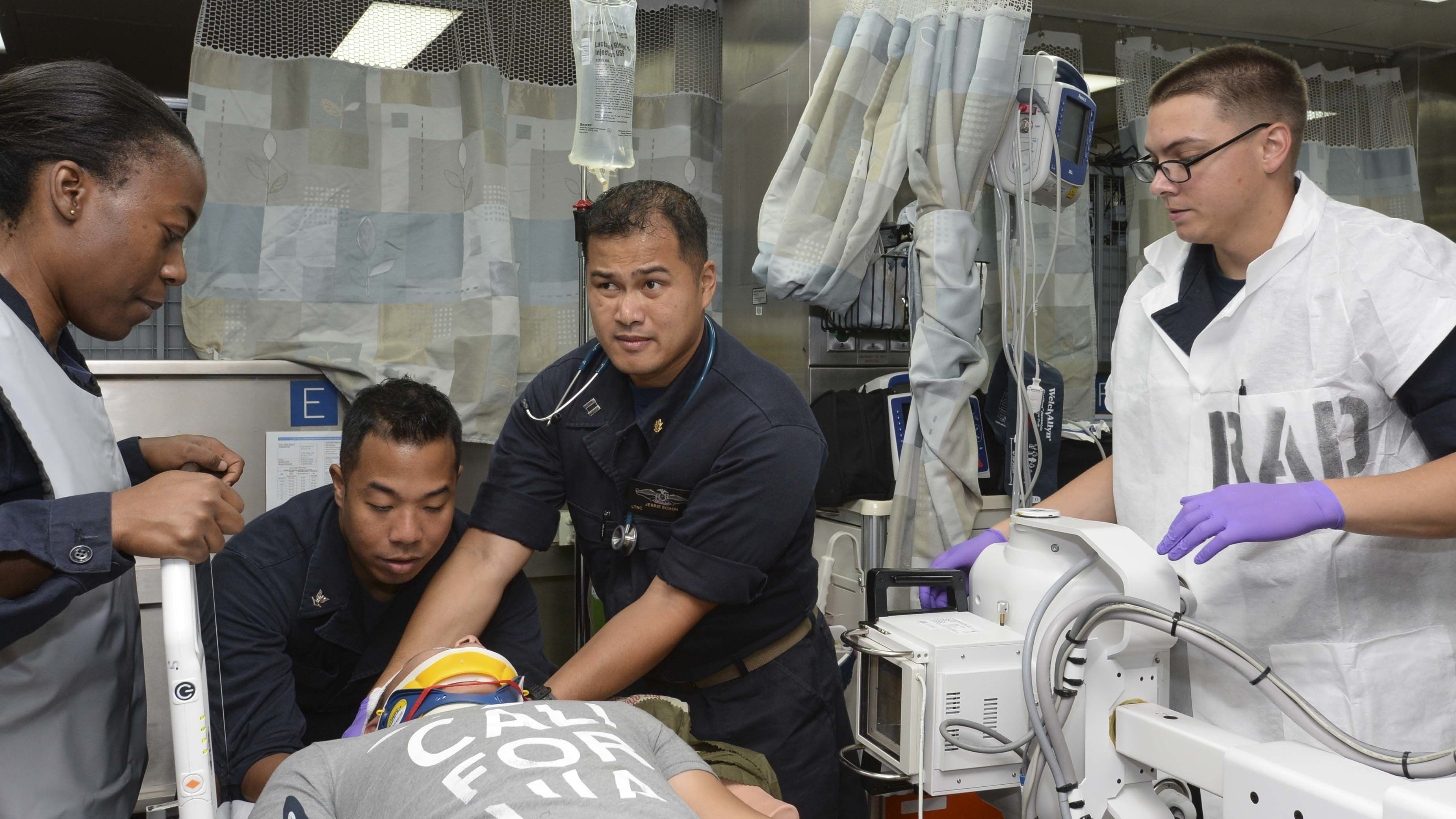 The image shows a group of four people standing around a man laying on a gurney in a hospital room....
