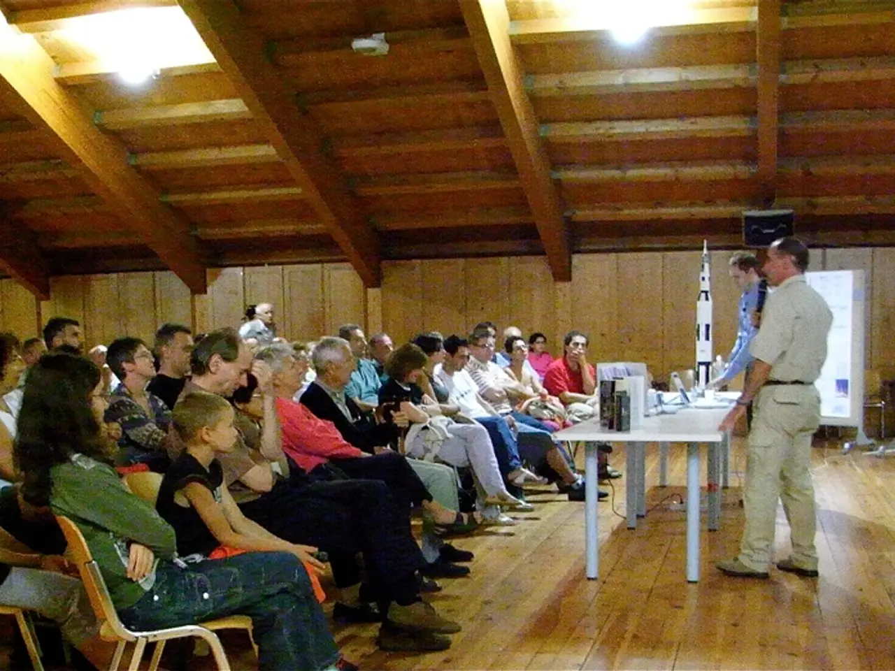 In this image there are group of people sitting in the hall and listening to the person who is...