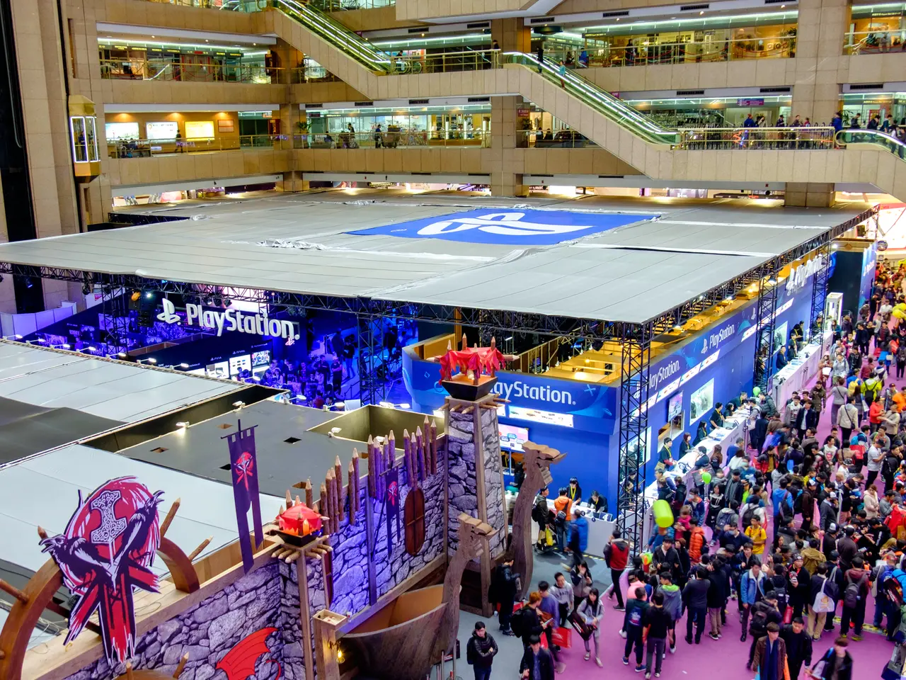 The image shows a large crowd of people standing in a large building with escalators, railings,...