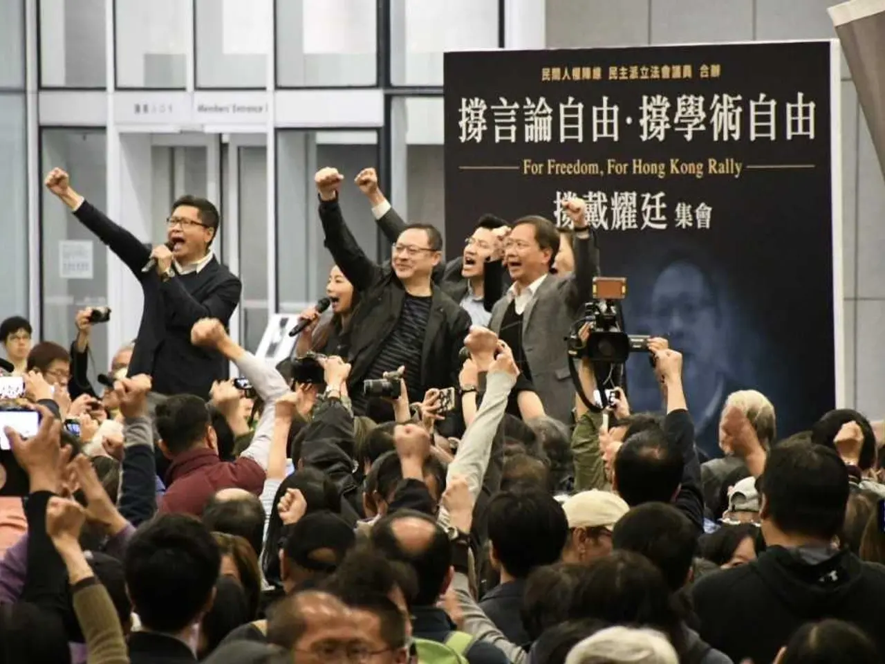 The image shows a group of people standing in front of a crowd of people, some of whom are holding...