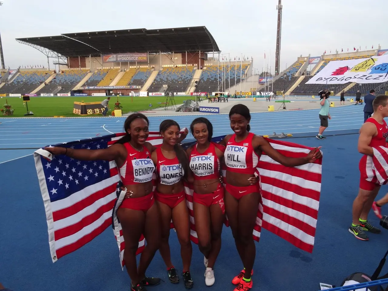 The image shows a group of four women standing next to each other on a track, smiling and holding...