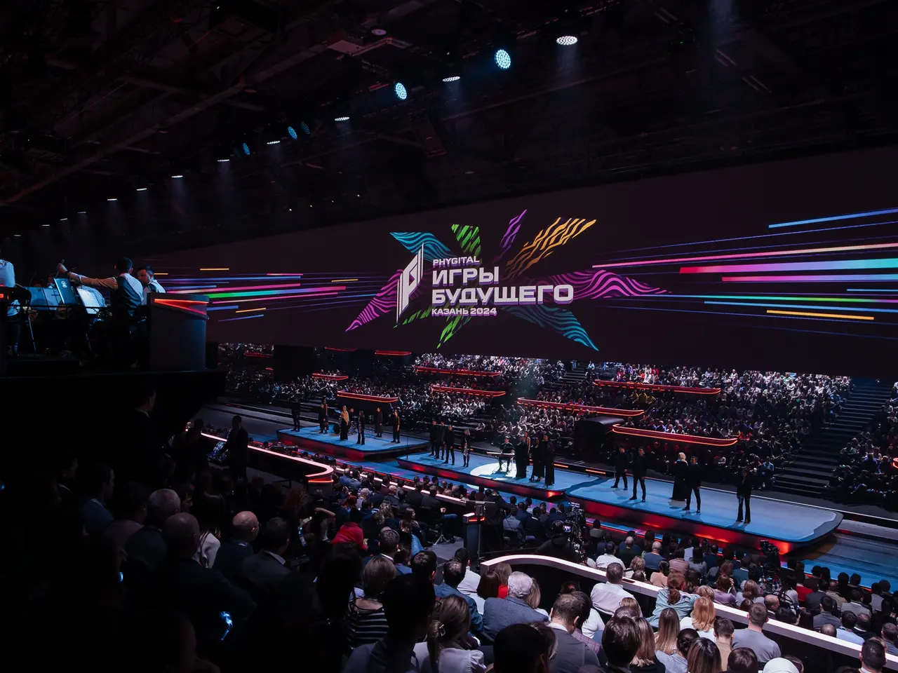 The image shows a large crowd of people sitting in front of a stage, with a few people standing on...