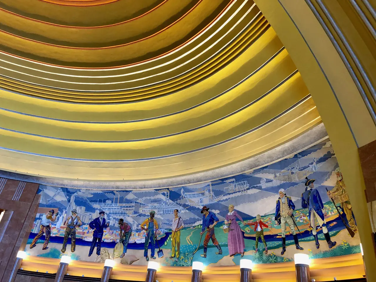 The image shows a large mural on the ceiling of a building, depicting people, buildings, and other...