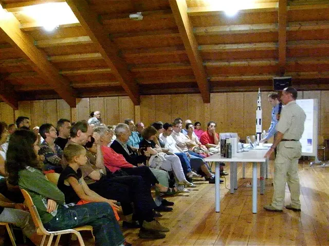 In this image there are group of people sitting in the hall and listening to the person who is...