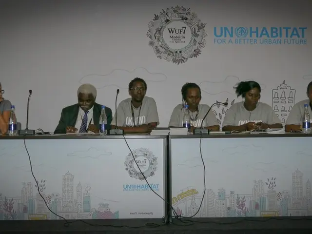 This image is clicked in a conference hall. There are six persons sitting in this image. To the...