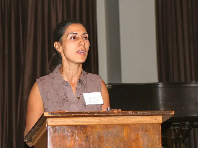 Here we can see a woman and there is a podium. In the background we can see curtains and a wall.