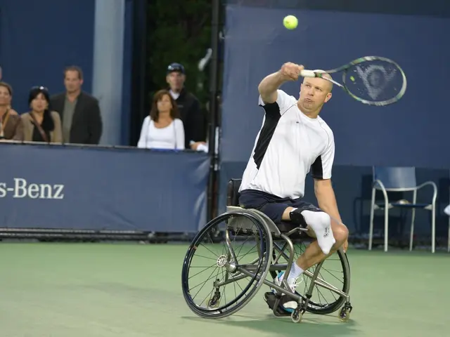 In this image there is a man sitting and running in a wheel chair playing a tennis racket game , at...