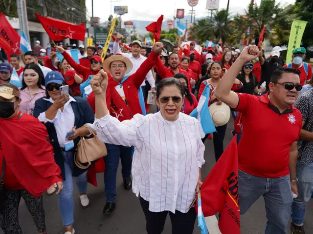The image shows a large group of people walking down a street, some of them holding flags and...