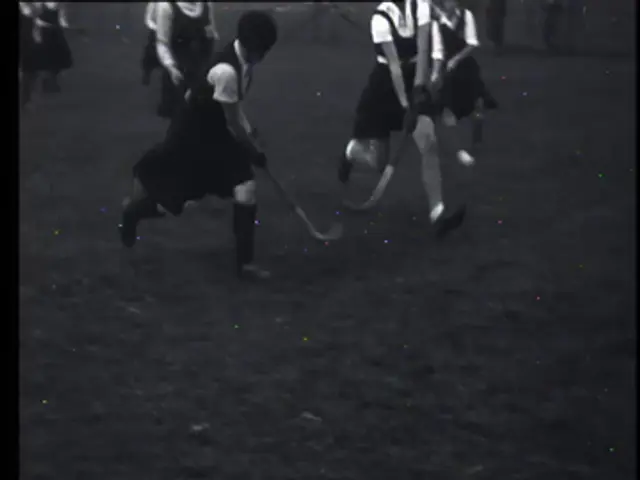 The image shows a group of young girls playing field hockey on a field, each of them holding a...